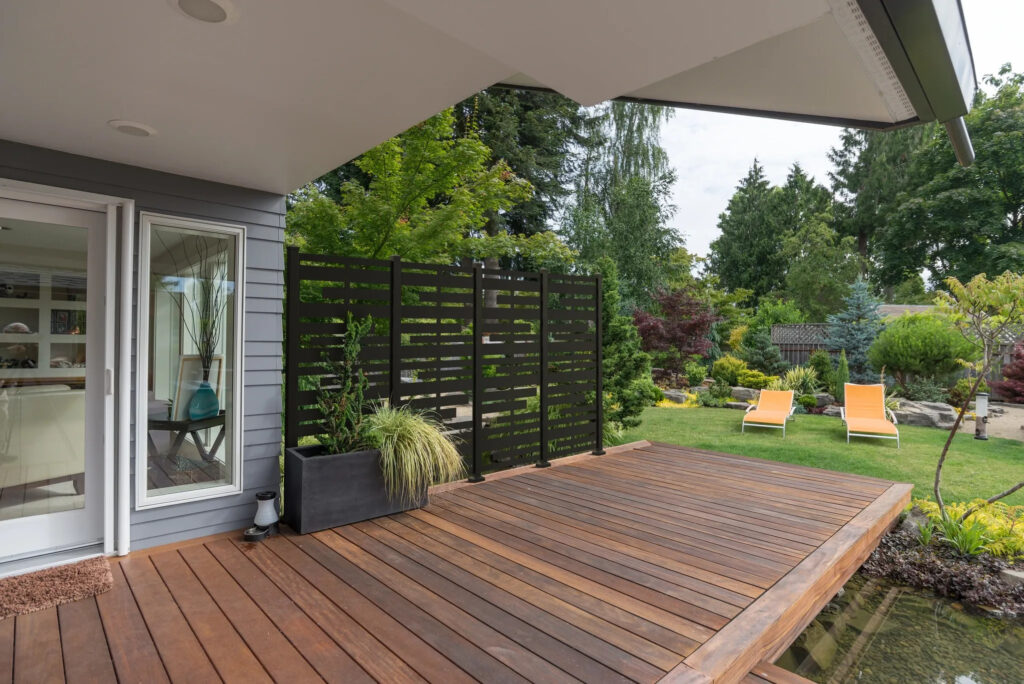 A perspective view of a contemporary Pacific Northwest home with a deck bridging a pond that leads to a pair of modern yellow loungers in a landscaped yard. (A perspective view of a contemporary Pacific Northwest home with a deck bridging a pond that